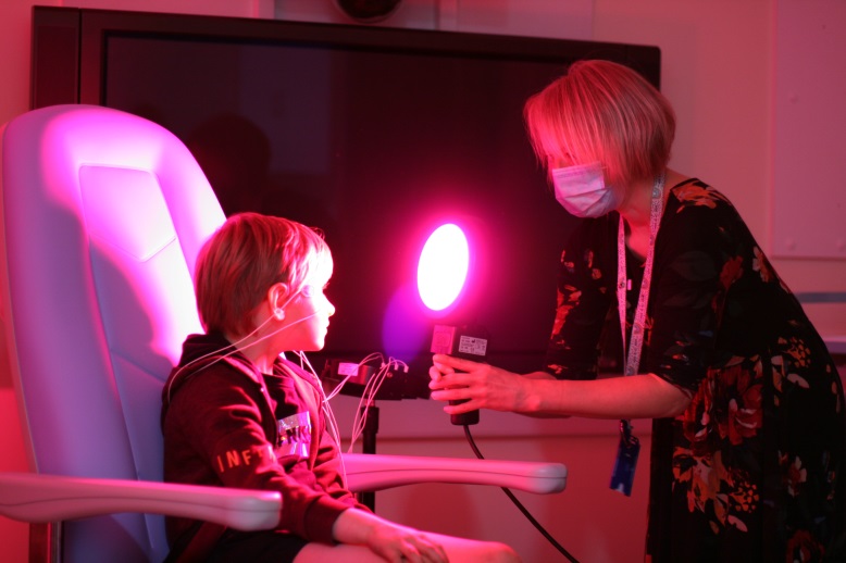A child tries out a device to help with eye tests, the paddle glows read and is held by a member of hospital staff in a face mask