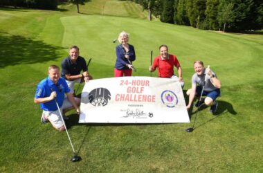 Lady Elsie and group holding 24 hour golf challenge poster on putting green.